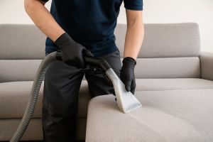 Ein Reinigungstechniker mit schwarzen Einmalhandschuhen säubert mit einem Extraktionsgerät die Sitzfläche eines hellgrauen Sofas – professionelle Polsterreinigung durch Herdegen Gebäudeservice GmbH.